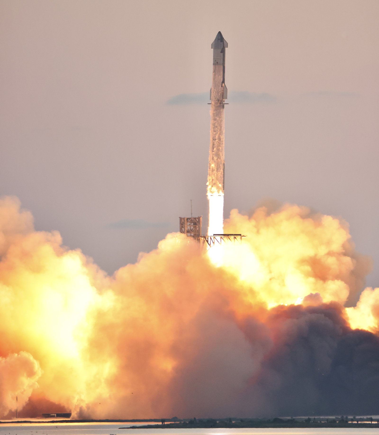 Starship full stack lifting off at dawn beside Mechazilla on Flight 5