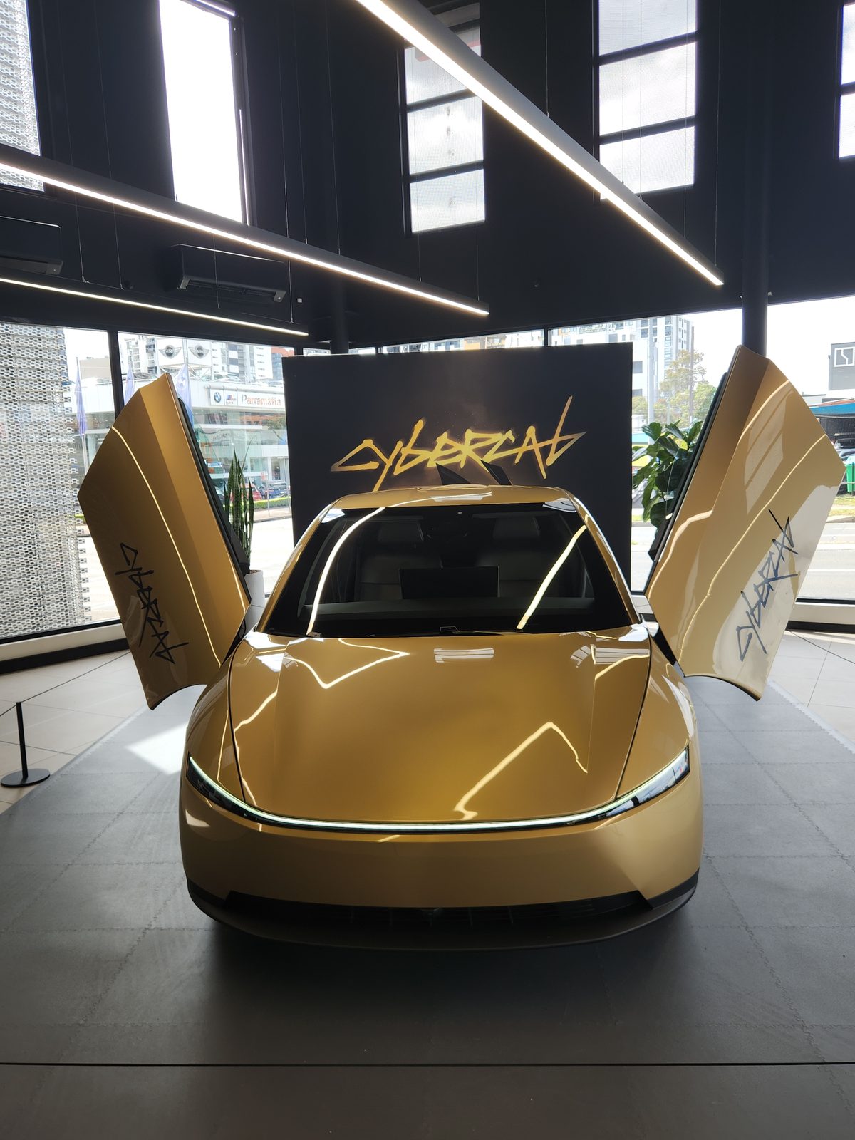 Front high view of gold Cybercab with both butterfly doors up and CYBERCAB wall behind