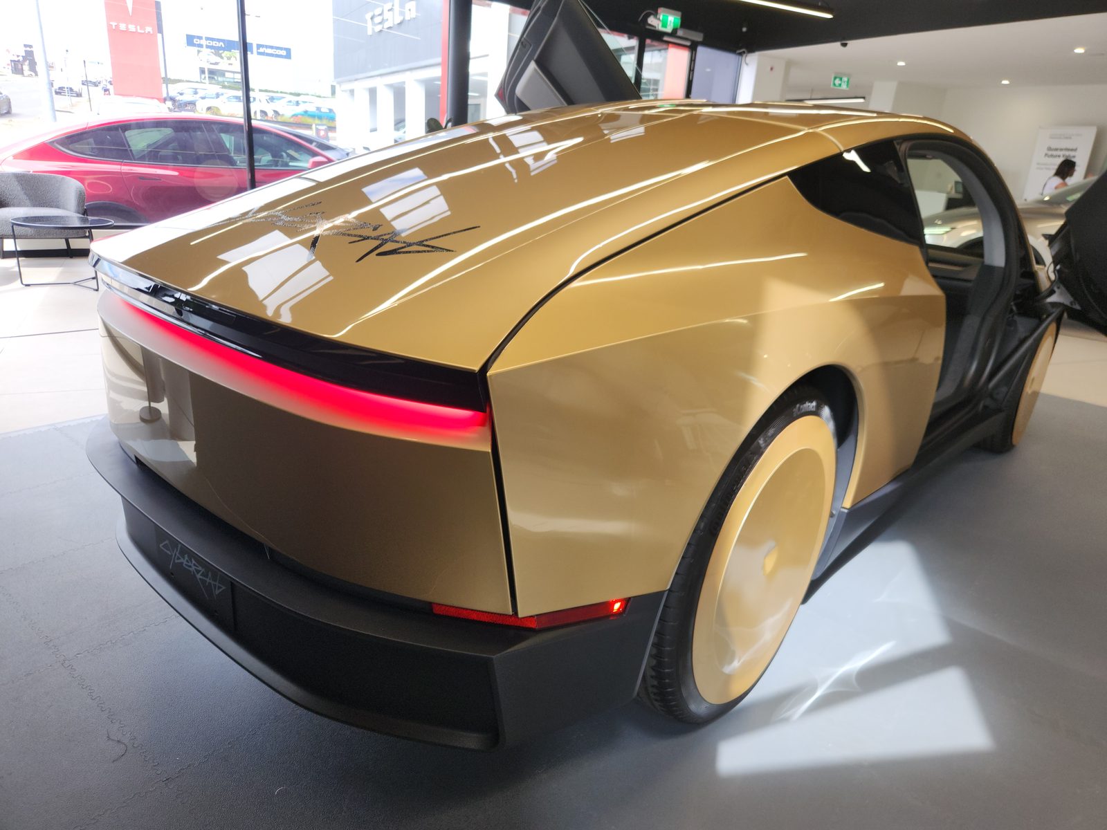 Rear three-quarter of gold Cybercab with door up and full-width red light bar