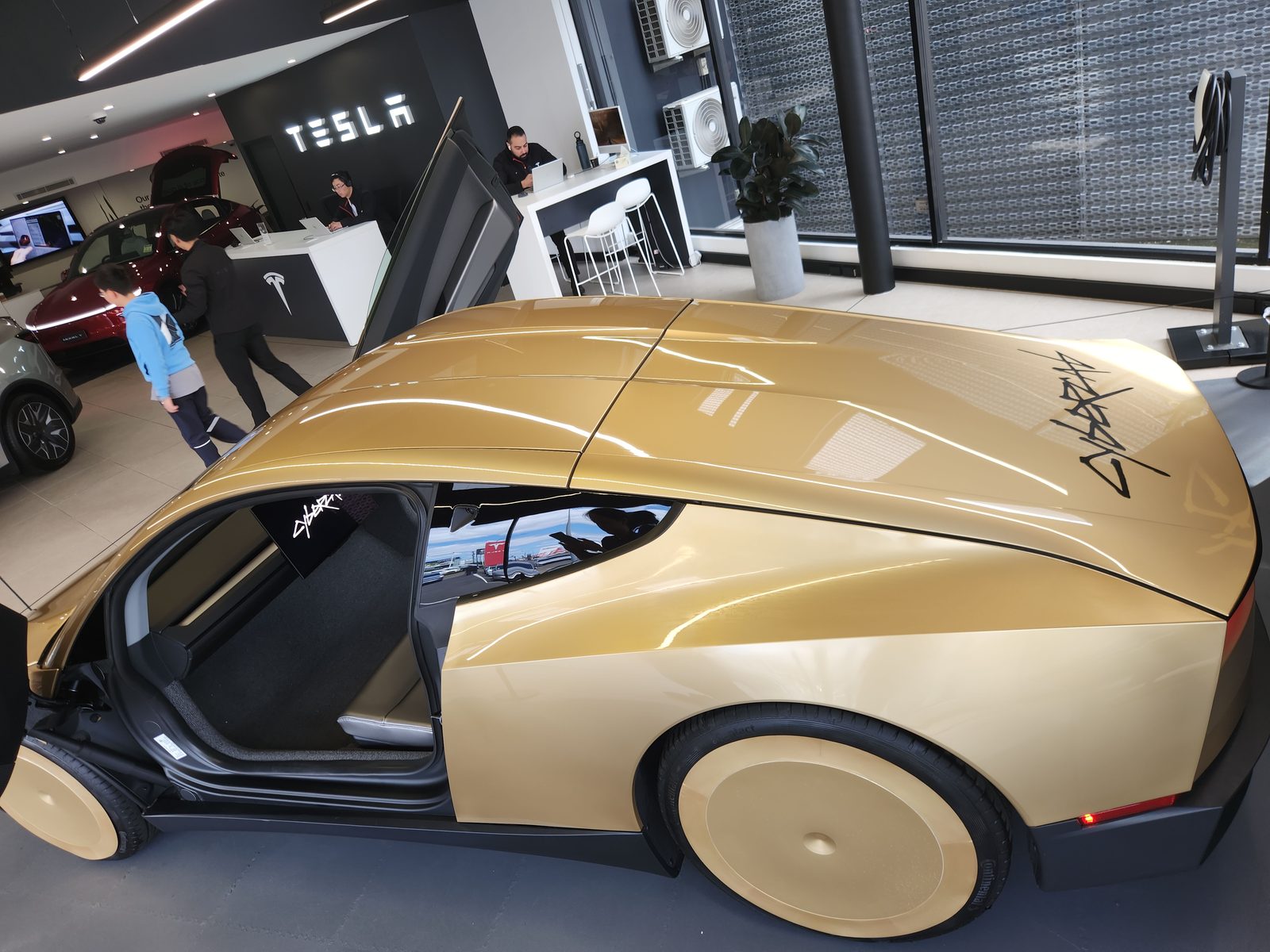 High side view of gold Cybercab with door open, TESLA wall and Cybercab logo on rear