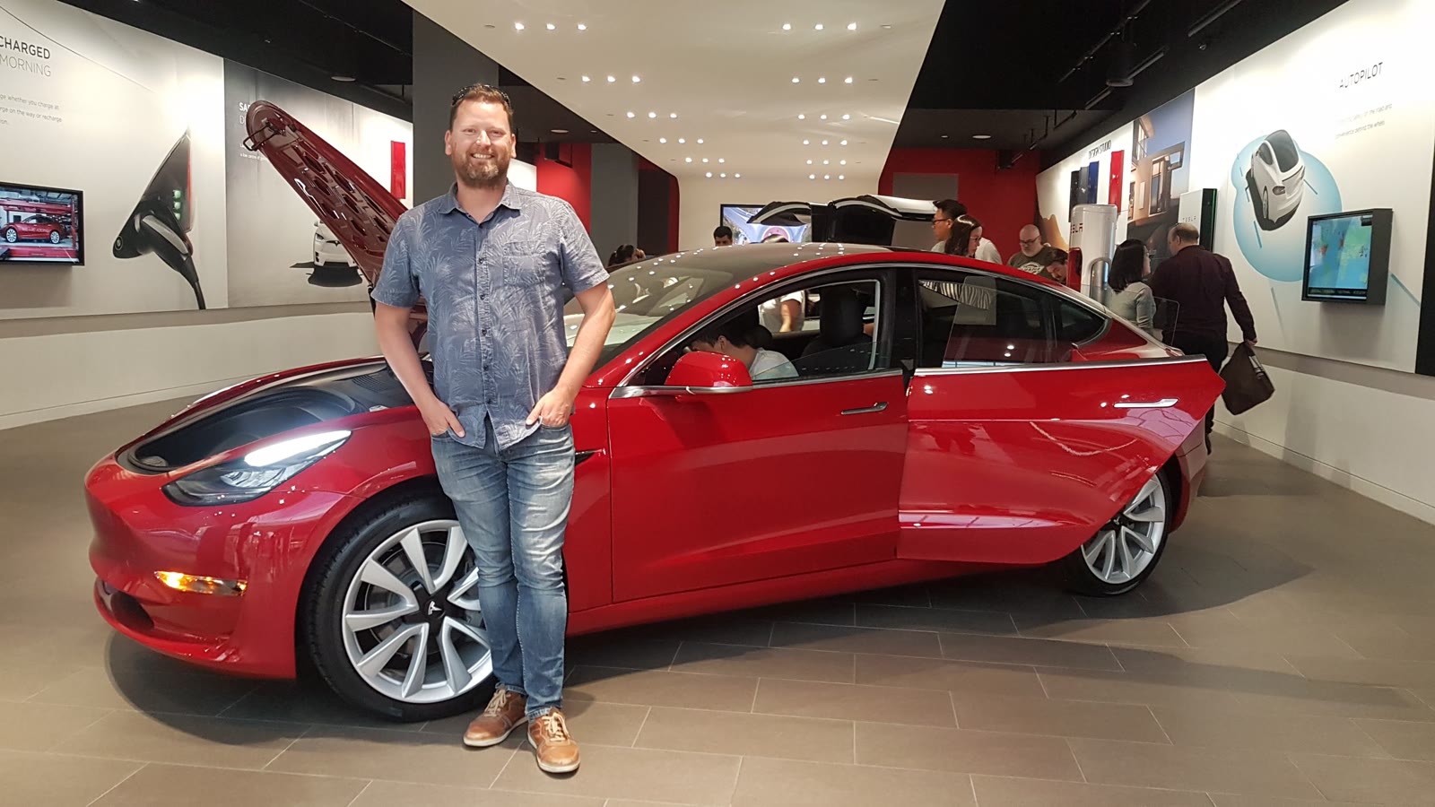 David standing beside a red Model 3 with the frunk open in a Tesla showroom